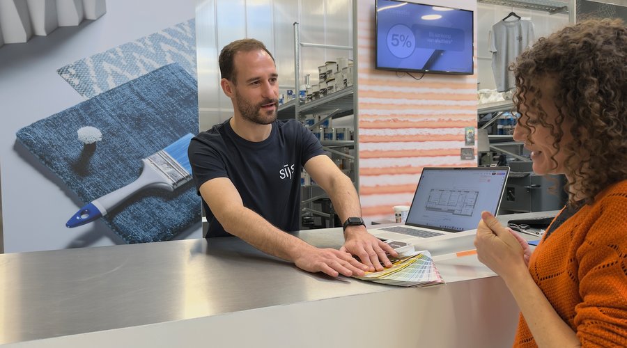 Verfspecialist geeft persoonlijk verfadvies aan een klant aan de toonbank, met kleurstalen en een plattegrond op de laptop