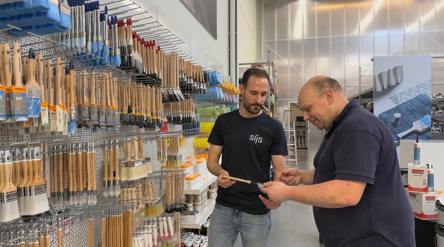 Sijs-medewerker helpt een klant bij het kiezen van een penseel voor schilderwerk, voor een uitgebreide wand vol kwasten en rollers